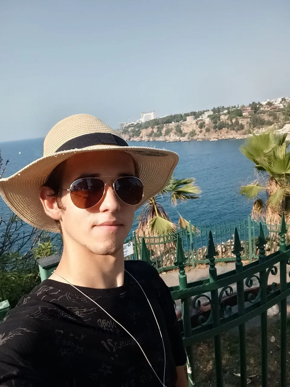 Mateusz Muszarski in a straw hat and sunglasses, the sea and a rocky coast behind him