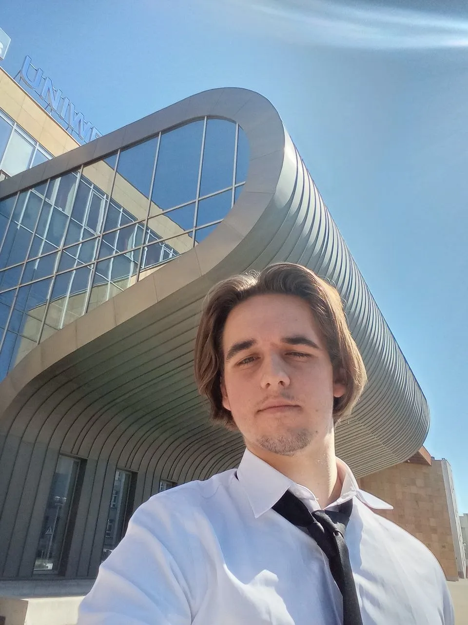 Mateusz Muszarski in a white shirt and black tie in front of a curved glass university building