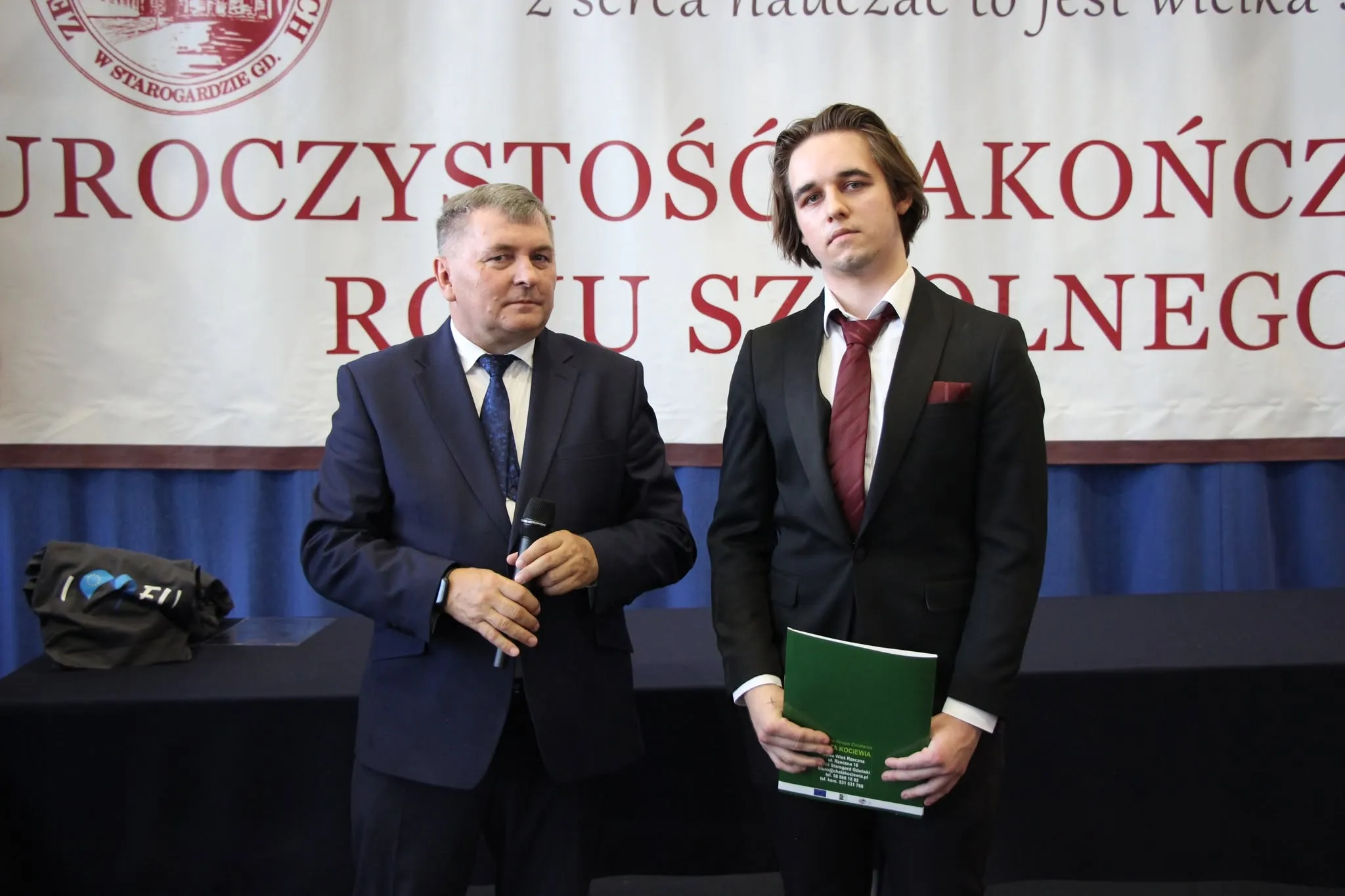 Mateusz Muszarski in a dark suit and red tie next to his school's principal, under an end-of-school-year banner